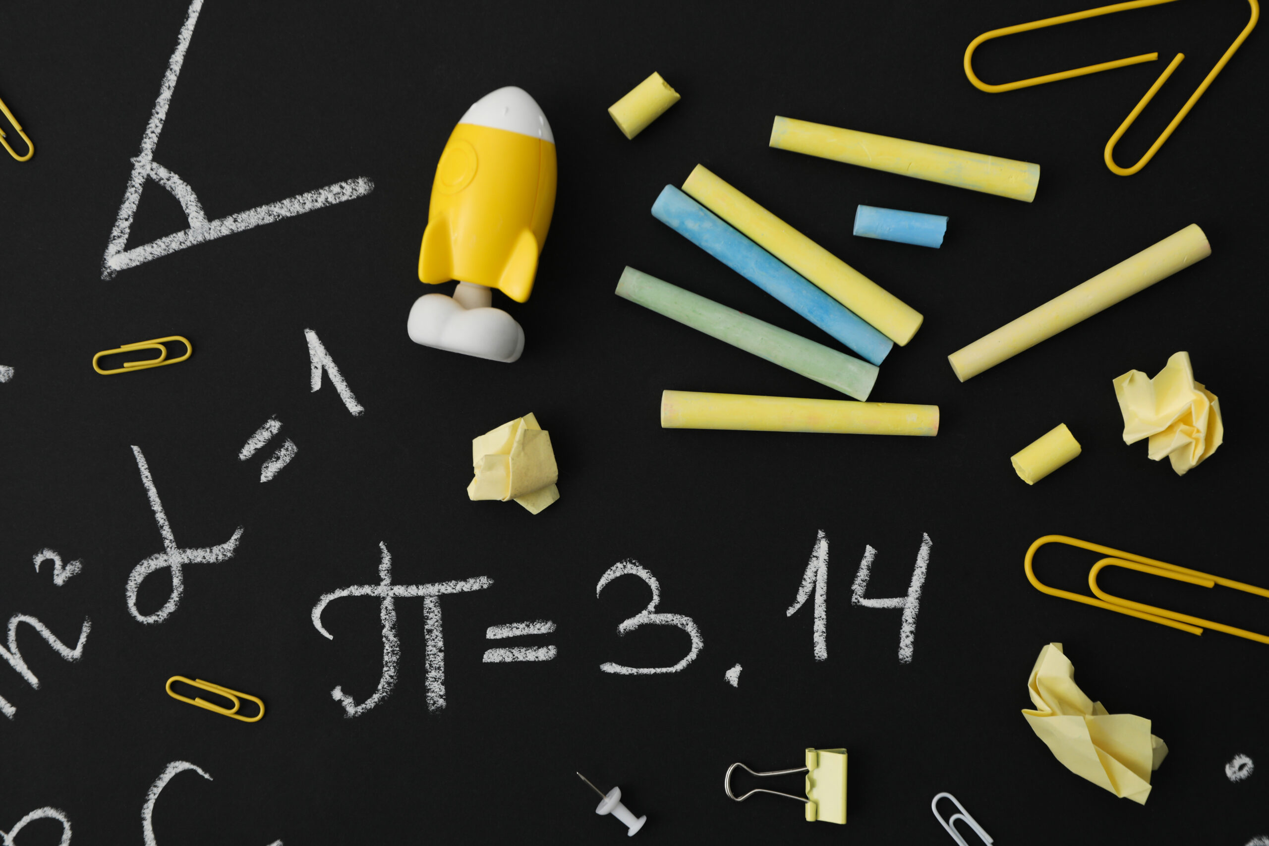 Black chalkboard with pencils and school materials spread across it. Pi number, 3.14 is written in chalk across the middle